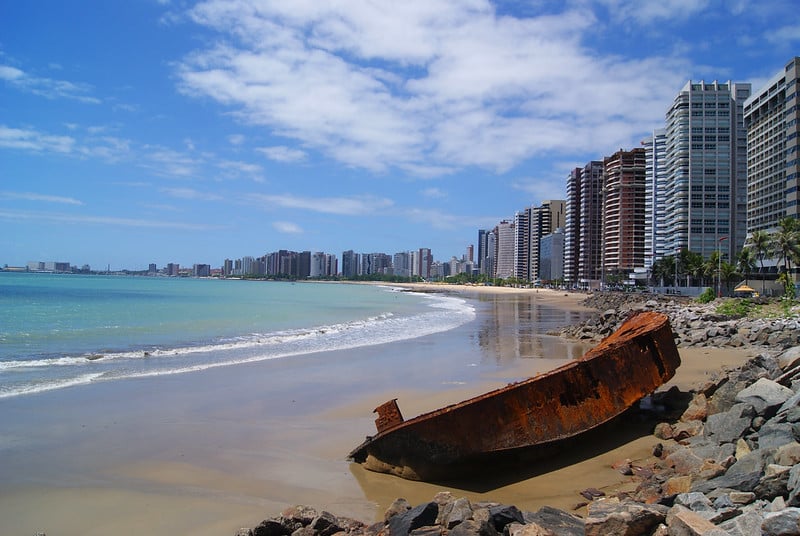 Foto beira-mar na praia de Iracema, em Fortaeza. Há um barco encalhado na areia à direita, e no fundo há vários prédios. Na esquerda está o mar. O céu é azul e com algumas nuvens.